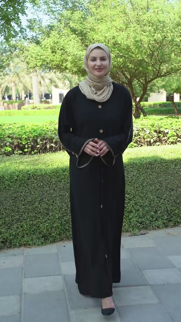 Schwarze Abaya mit beigefarbenen Paspeln und Zierknöpfen vorne und an den Ärmeln in Schwarz.